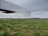 a mirrored structure in the middle of a grassy field overlooking a river with buildings in the background