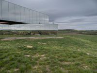 a mirrored structure in the middle of a grassy field overlooking a river with buildings in the background