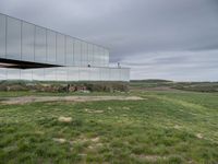 a mirrored structure in the middle of a grassy field overlooking a river with buildings in the background