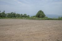 an empty parking lot with a dirt surface and trees in the background, overlooking a distant hillside