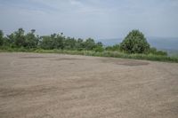 an empty parking lot with a dirt surface and trees in the background, overlooking a distant hillside