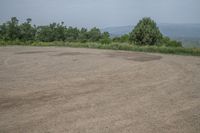 an empty parking lot with a dirt surface and trees in the background, overlooking a distant hillside