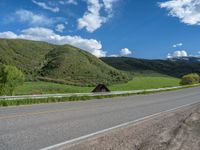 the empty road is going through a valley with mountains and grass in the background and two houses on both sides