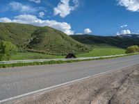 the empty road is going through a valley with mountains and grass in the background and two houses on both sides