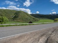 the empty road is going through a valley with mountains and grass in the background and two houses on both sides