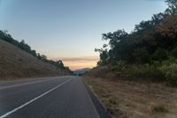 a car driving down a highway with trees around it at sunset at dusk time against a mountain wall