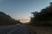 a car driving down a highway with trees around it at sunset at dusk time against a mountain wall