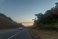 a car driving down a highway with trees around it at sunset at dusk time against a mountain wall