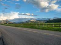 the road is empty during the day and has mountains in the distance as well as a fence