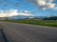 the road is empty during the day and has mountains in the distance as well as a fence