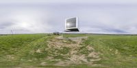 the view of an old tv standing on a grassy hill in a large field, with clouds hovering over the hill