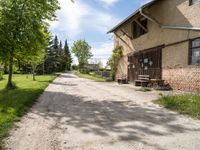 dirt road in front of a building with trees and grass around it, near by a bench