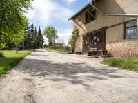 dirt road in front of a building with trees and grass around it, near by a bench