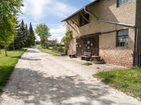 dirt road in front of a building with trees and grass around it, near by a bench