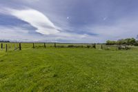 the view from behind a wooden gate into a large grassy field with clouds in the sky