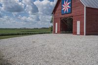 a barn on top of a gravel road next to grass and trees in front of a blue sky