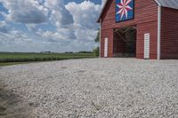 a barn on top of a gravel road next to grass and trees in front of a blue sky