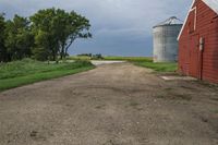 a dirt road going toward two barns in rural country setting with storm clouds overhead above