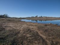 a big pond is surrounded by dirt in the field under a blue sky there are small trees on the hill