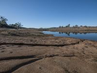 a big pond is surrounded by dirt in the field under a blue sky there are small trees on the hill