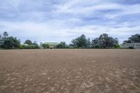 a field with lots of dirt and a large house in the distance there are trees in the foreground