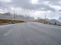 an empty road leads uphill towards mountains under cloudy skies over the horizon are power lines