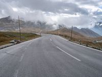 an empty road leads uphill towards mountains under cloudy skies over the horizon are power lines