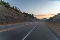 the road is next to some hills at sunset with a small tree line on the right side