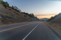 the road is next to some hills at sunset with a small tree line on the right side