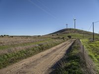 a dirt road runs alongside the wind turbines on a hill on an otherwise rural land