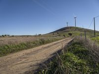 a dirt road runs alongside the wind turbines on a hill on an otherwise rural land