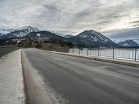 an open asphalt road leads to a body of water with snow capped mountains behind it