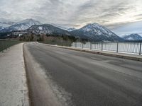 an open asphalt road leads to a body of water with snow capped mountains behind it