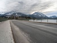 an open asphalt road leads to a body of water with snow capped mountains behind it