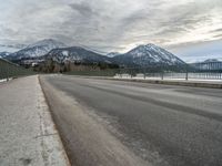 an open asphalt road leads to a body of water with snow capped mountains behind it