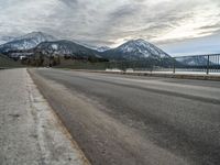 an open asphalt road leads to a body of water with snow capped mountains behind it