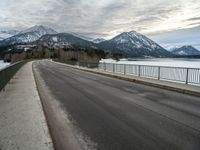 an open asphalt road leads to a body of water with snow capped mountains behind it