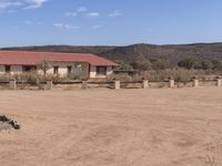 a dirt road with a fence and building in the background with a red roof and several concrete pillars