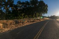 an asphalt road in the evening with the sun setting over it and trees behind it