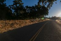 an asphalt road in the evening with the sun setting over it and trees behind it