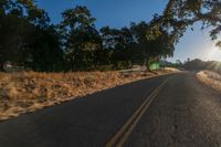 an asphalt road in the evening with the sun setting over it and trees behind it