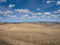 an empty field full of dry grass with clouds in the blue sky above it and hills on the far side