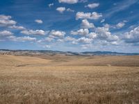 an empty field full of dry grass with clouds in the blue sky above it and hills on the far side