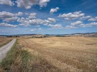 an empty field full of dry grass with clouds in the blue sky above it and hills on the far side