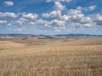 an empty field full of dry grass with clouds in the blue sky above it and hills on the far side