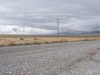 gravel road with power lines in the distance next to a dry field and mountains with a lot of clouds