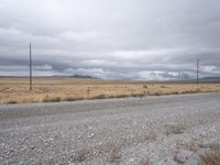 gravel road with power lines in the distance next to a dry field and mountains with a lot of clouds