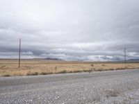 gravel road with power lines in the distance next to a dry field and mountains with a lot of clouds