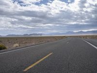 a road in a desert with mountains on both sides of it and clouds overhead and an area full of arid land below