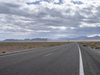 a road in a desert with mountains on both sides of it and clouds overhead and an area full of arid land below